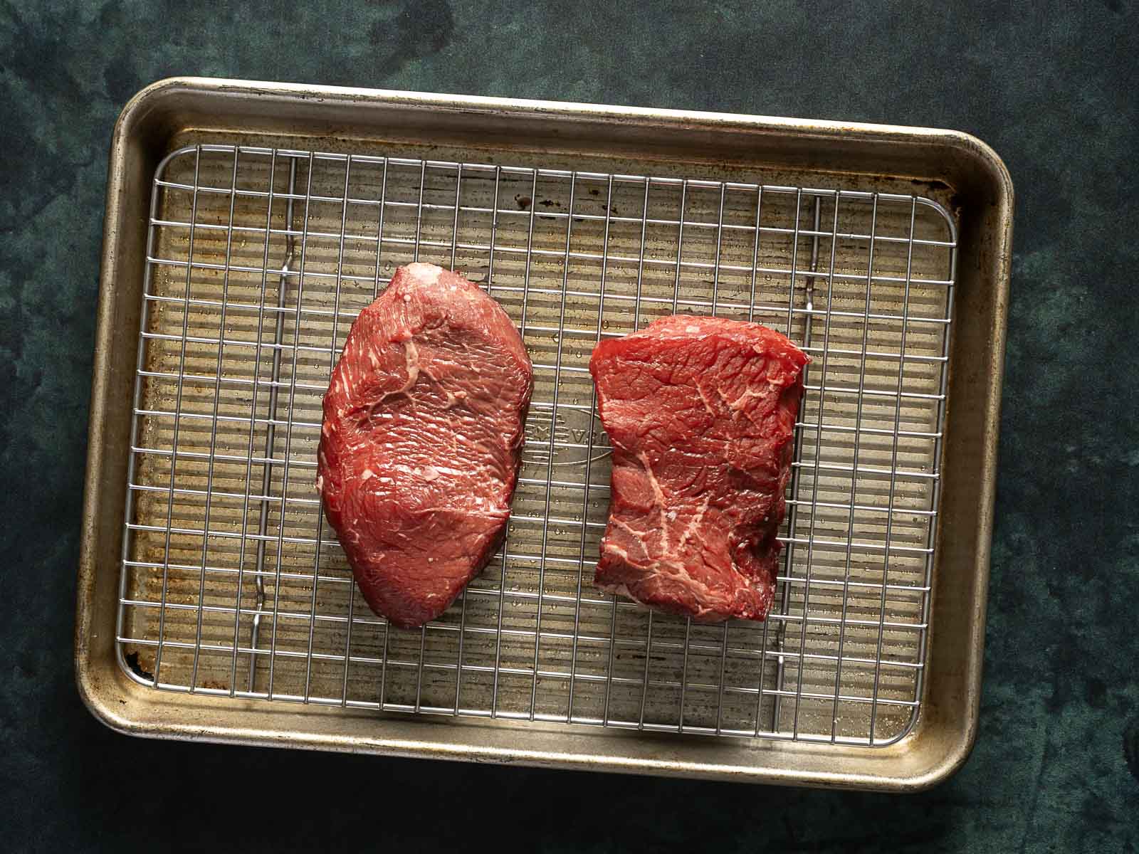 Two raw steaks on a wire rack placed in a metal baking tray, set against a dark surface.