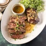 Sliced and whole steak topped with herbs and garlic, served with a side of microgreens and a small bowl of sauce on a white oval plate.