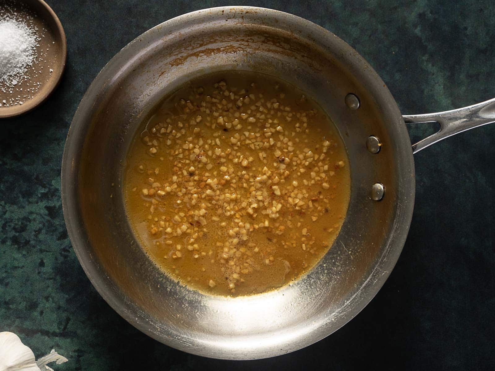 A stainless steel pan containing a mixture of finely chopped garlic and oil sits on a dark green countertop. A small bowl of salt is partially visible in the corner.