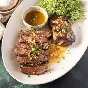 Sliced and whole steak topped with herbs and garlic, served with a side of microgreens and a small bowl of sauce on a white oval plate.