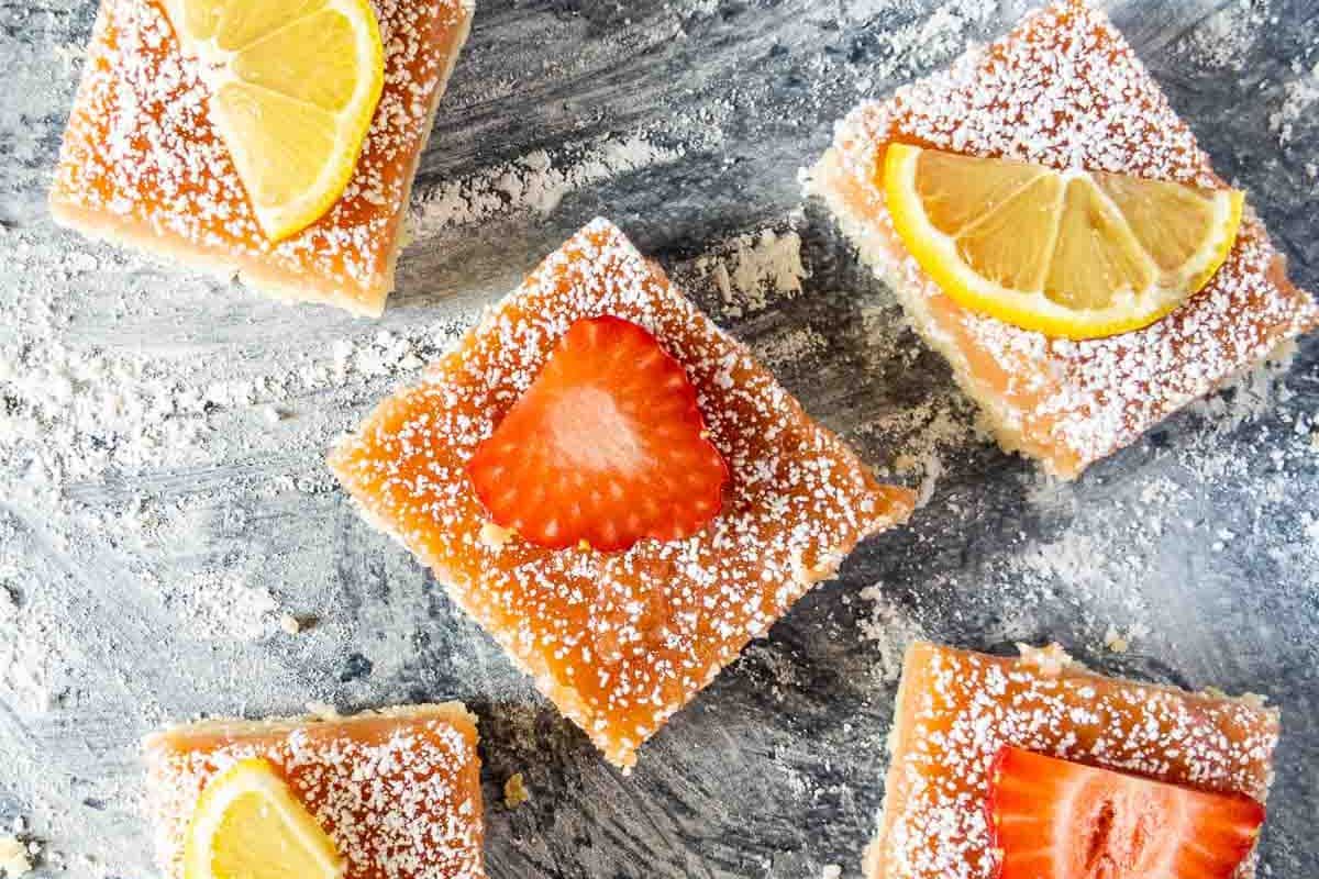 Square lemon bars, a delightful Easter dessert, are topped with powdered sugar, lemon slices, and strawberry pieces. These treats, arranged on a textured surface, are also gluten-free for everyone to enjoy.