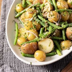 A platter of baby potatoes and green beans mixed with herbs and seasoning on a white dish, set on a textured gray cloth.