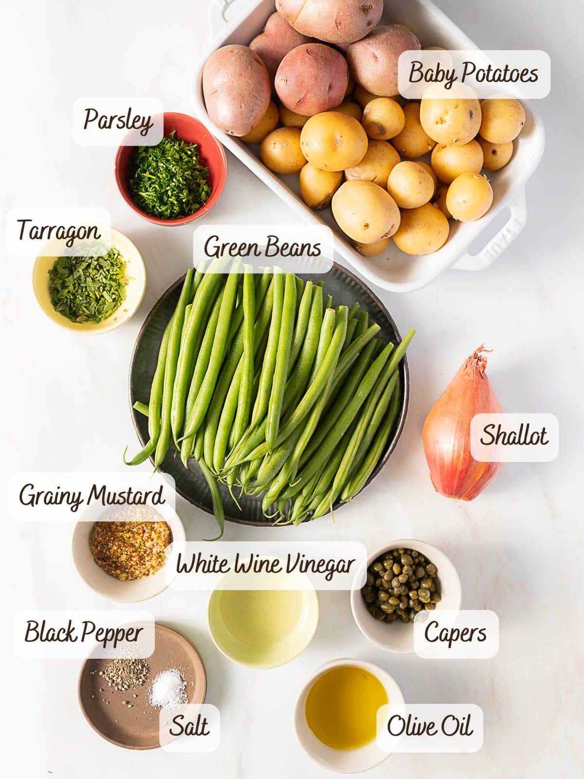 A display of labeled ingredients for a green bean potato salad, featuring baby potatoes, green beans, shallot, parsley, tarragon, white wine vinegar, grainy mustard, olive oil, salt, black pepper, and capers.