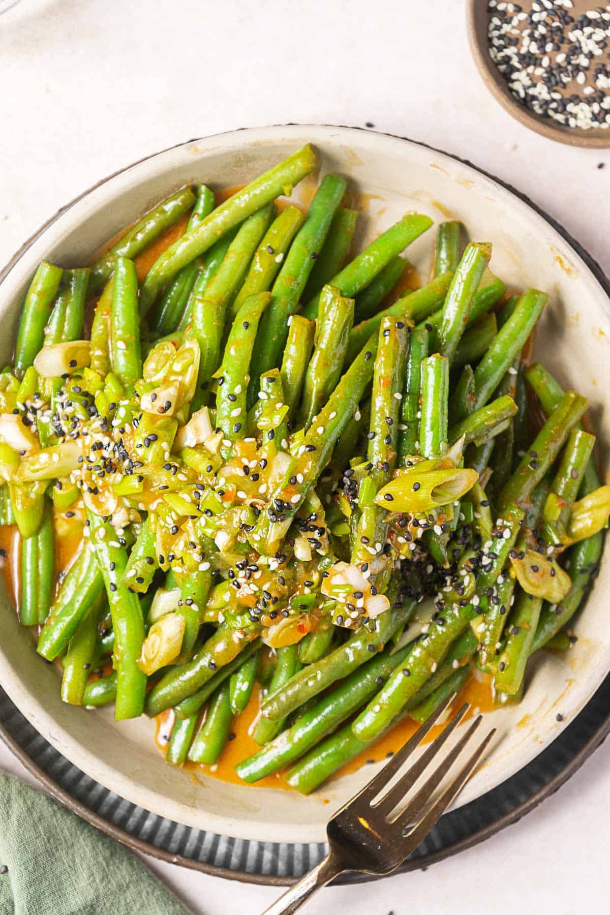 A plate of cooked green beans topped with sliced green onions and a sprinkle of sesame seeds, with a fork placed beside the plate.