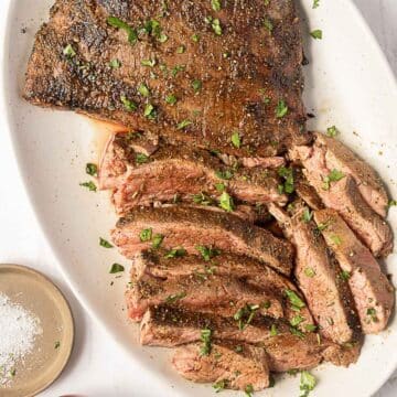 Sliced grilled flank steak served on a white platter, garnished with chopped herbs. Small dish of coarse salt is visible nearby.