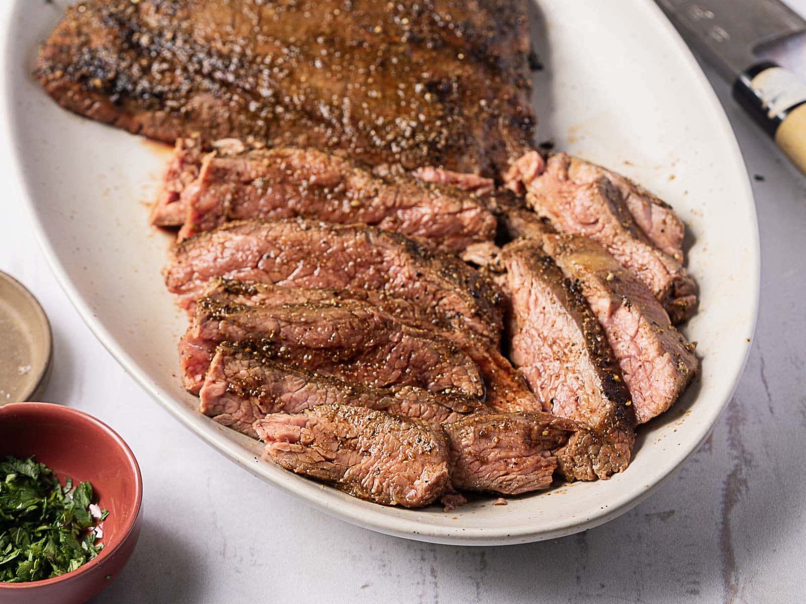 A white oval plate with sliced grilled steak, seasoned with pepper, displayed next to a small bowl of chopped herbs and a kitchen knife.