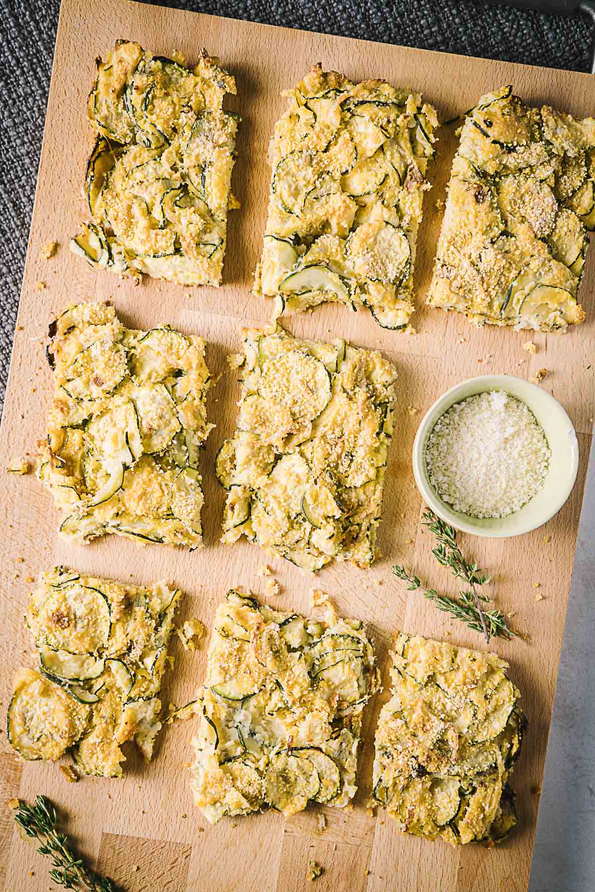 Eight squares of zucchini bread with a bowl of grated cheese and herbs on a wooden board.