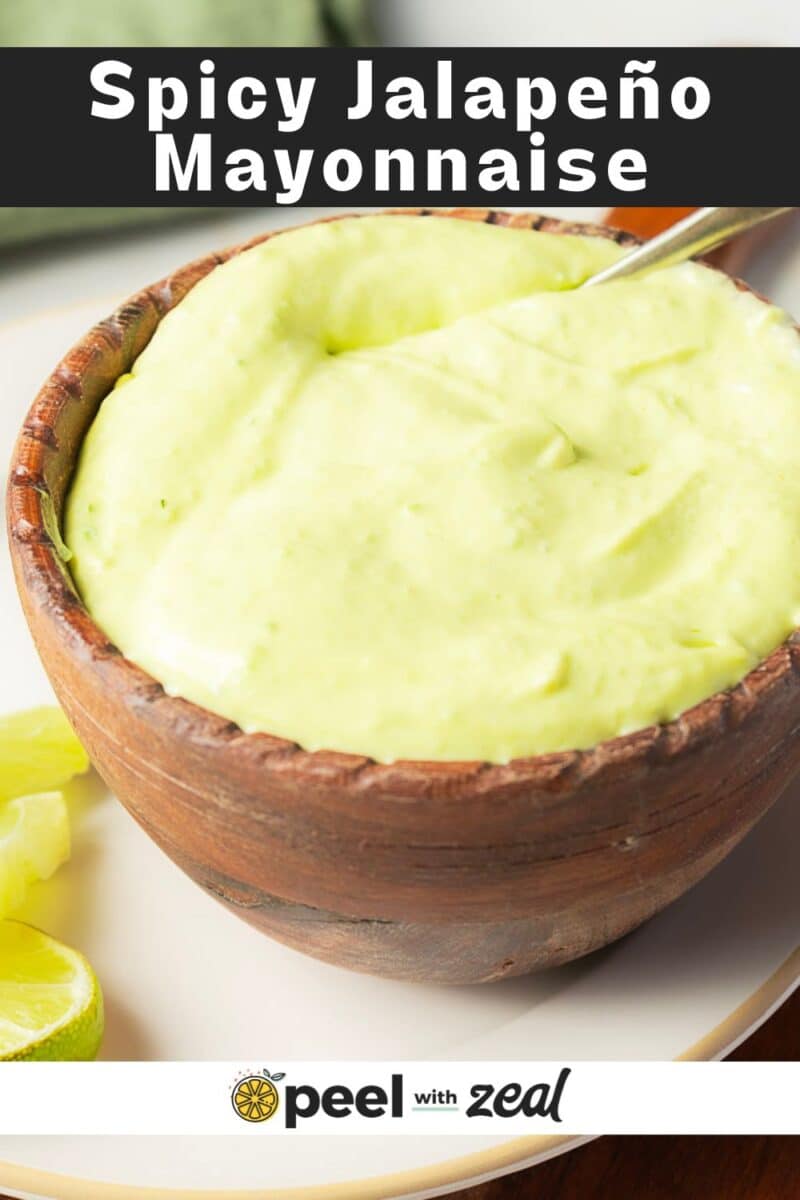 A wooden bowl filled with light green jalapeno mayo and a spoon sits next to fresh lime wedges on a plate.