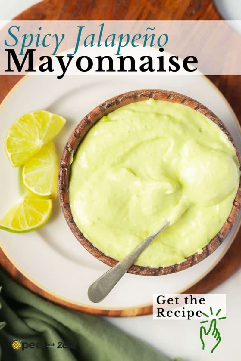 A bowl of creamy light green jalapeno mayo with a spoon, served on a plate with lime wedges. Text reads: "Spicy Jalapeño Mayonnaise. Get the Recipe.