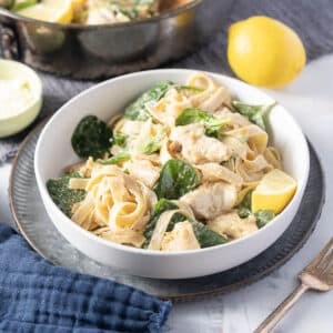 A bowl of fettuccine pasta with creamy sauce, spinach, and chicken pieces. A lemon wedge is on the side. Pan and lemon are in the background on a marble surface.
