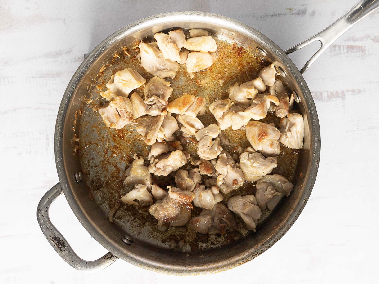 Chunks of chicken cooking in a stainless steel pan on a white surface.