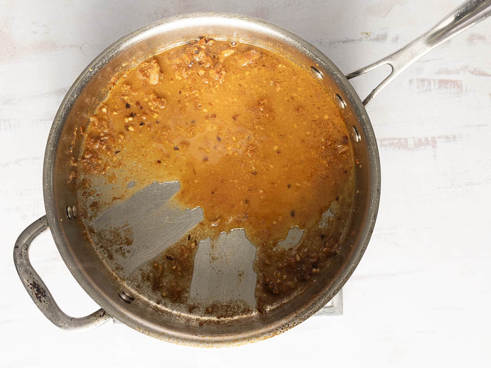 Partial view of a metal pan with a brown sauce and bits of meat, showing an area scraped clean, sitting on a white surface.