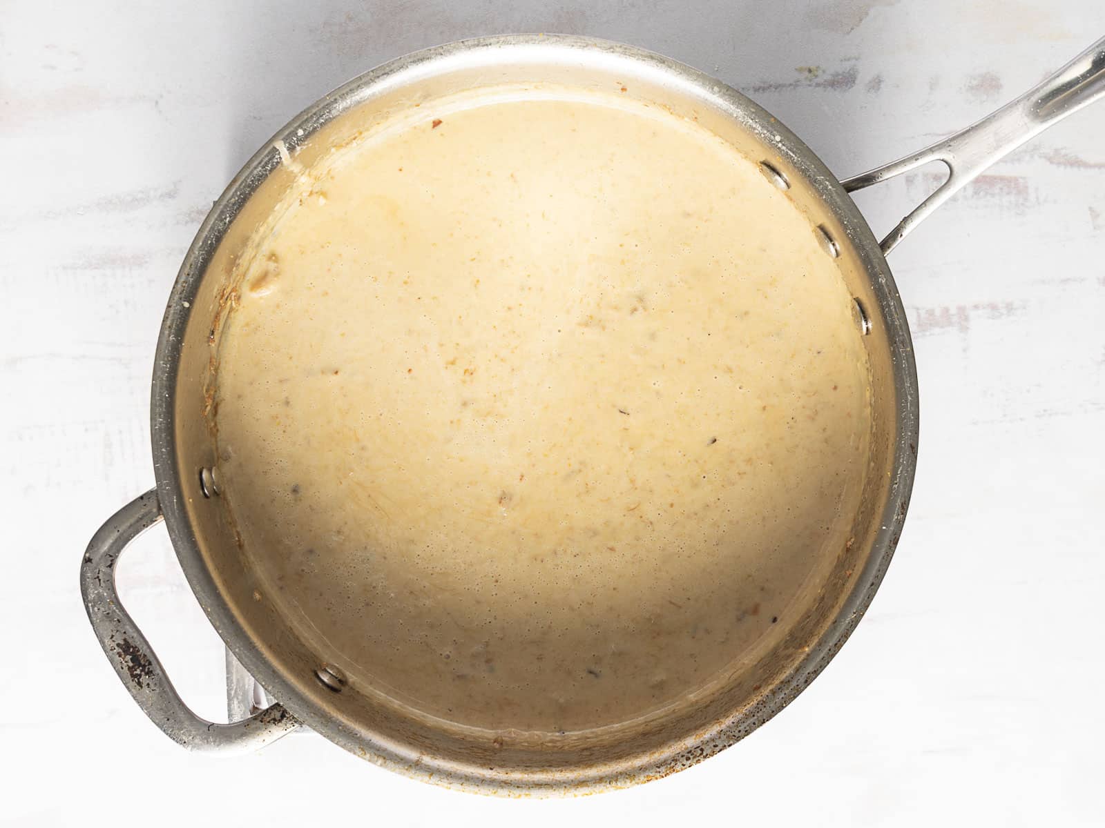 A creamy beige sauce simmering in a metal saucepan on a light-colored surface.