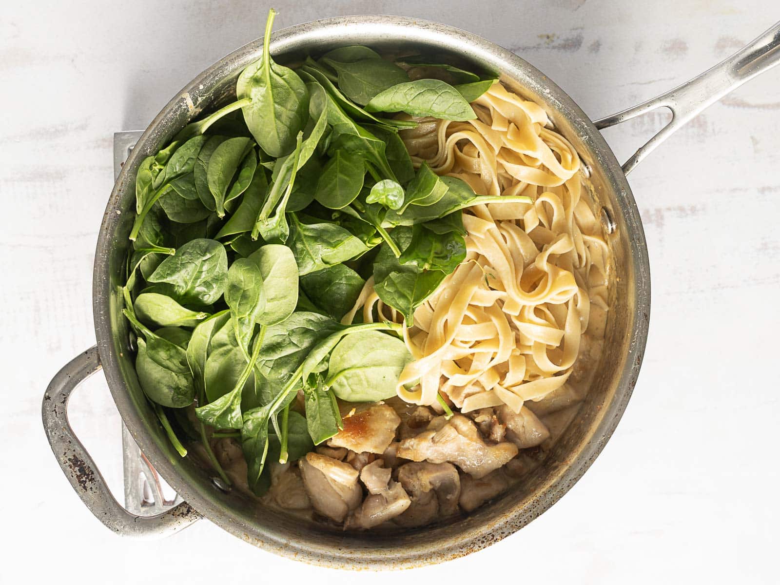 A pot containing raw spinach, cooked pasta, and chicken pieces on a white surface.