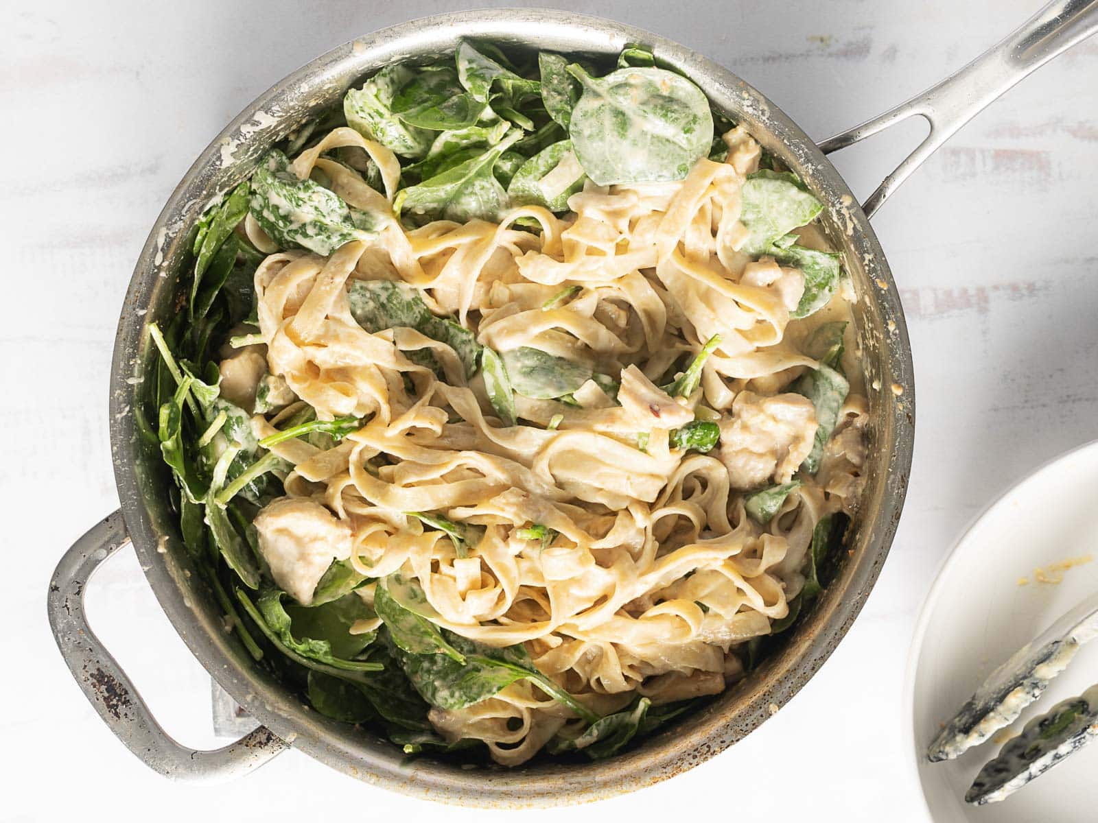 A pan filled with creamy pasta, mixed with spinach and chicken pieces, sits on a light surface.