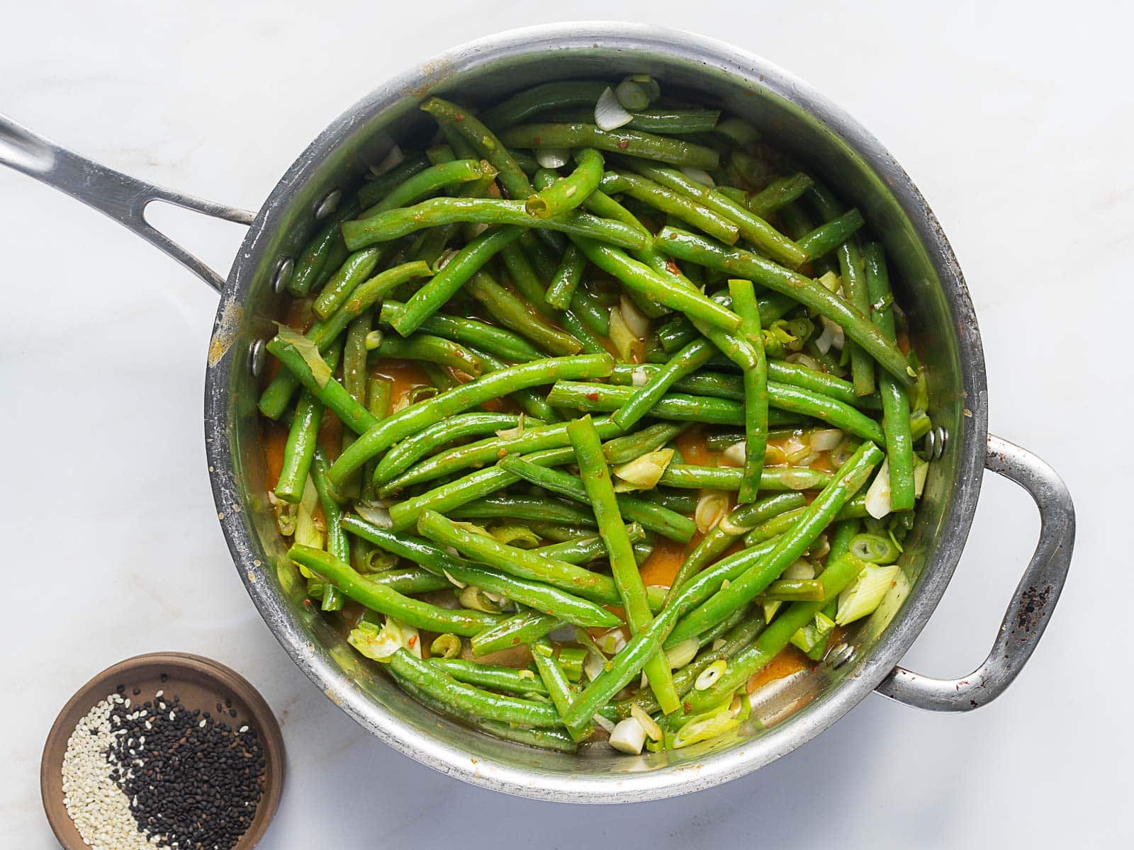 A stainless steel pan filled with cooked green beans, sliced garlic, and sauce sits on a white surface next to a small dish of black and white sesame seeds.