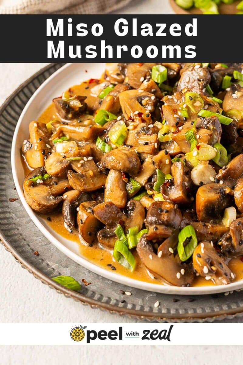 A plate of miso mushrooms glazed to perfection, garnished with chopped green onions and sesame seeds, served on a white dish with a black rim.