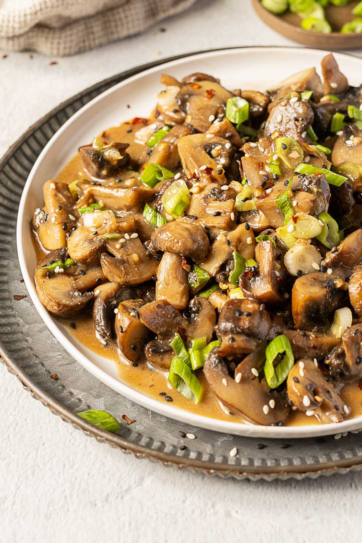A plate of sautéed mushrooms garnished with chopped green onions and sesame seeds, served on a white plate with a gray charger underneath.
