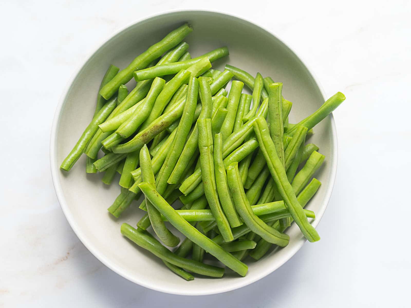 A white bowl filled with fresh, trimmed green beans on a light surface.