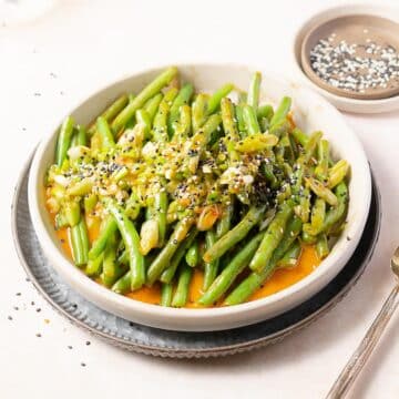 A bowl of cooked green beans garnished with sliced scallions and black and white sesame seeds, served on a white plate with a fork beside it.