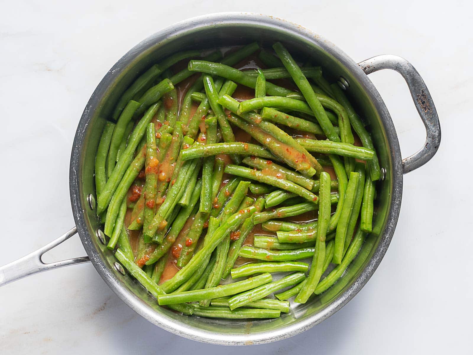 A stainless steel skillet containing cooked green beans in a light tomato-based sauce, photographed from above on a white surface.