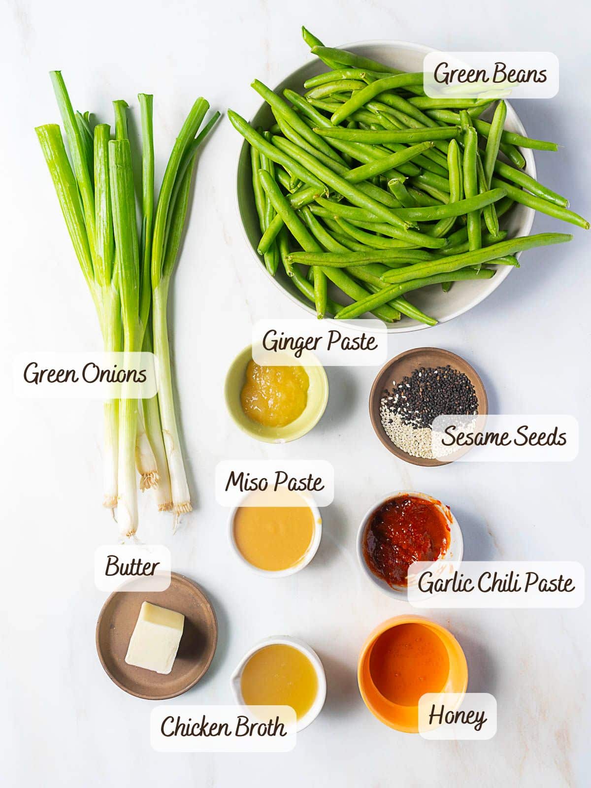 A bowl of miso green beans surrounded by small, labeled bowls of green onions, ginger paste, sesame seeds, butter, garlic chili paste, chicken broth, and honey.