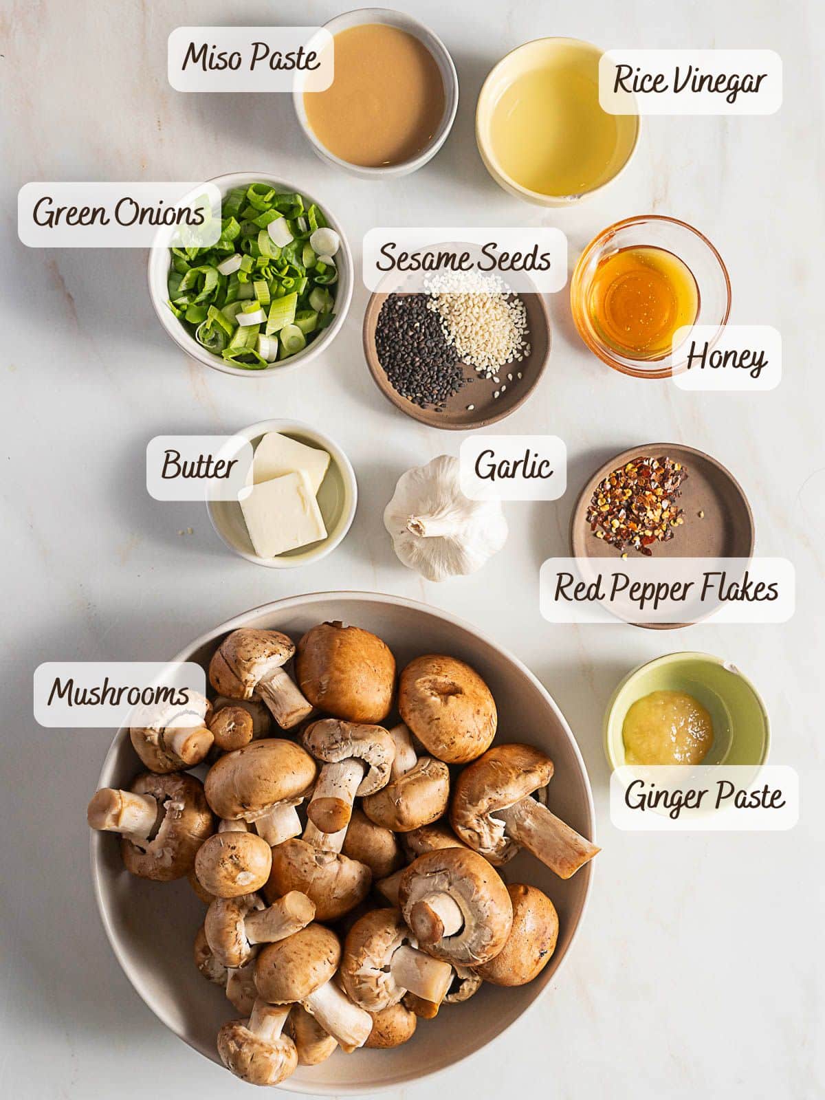 A bowl of miso mushrooms is surrounded by miso paste, rice vinegar, green onions, sesame seeds, honey, butter, garlic, red pepper flakes, and ginger paste in small dishes.