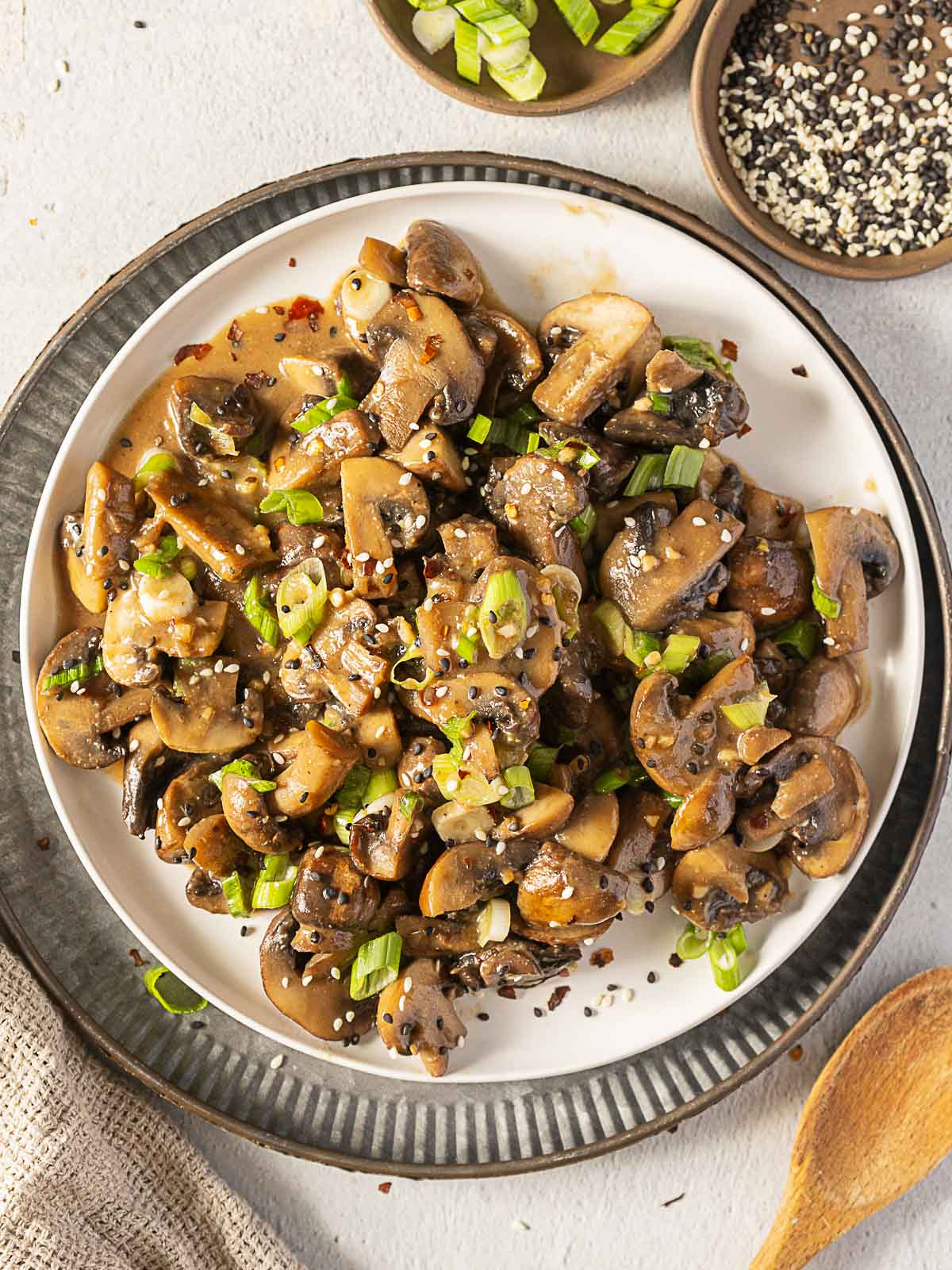 A plate of sautéed mushrooms garnished with chopped green onions and sesame seeds, served on a white plate with a dark rim.