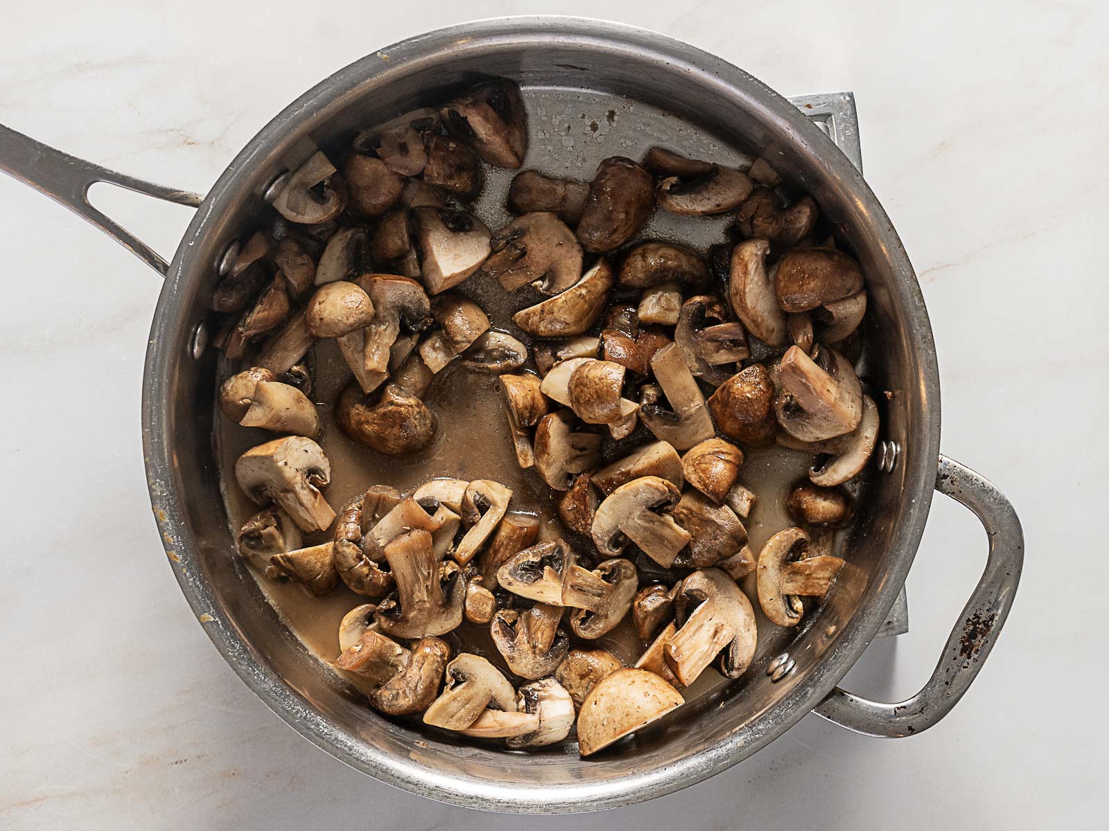 Sliced mushrooms cooking in a stainless steel pan on a white surface.