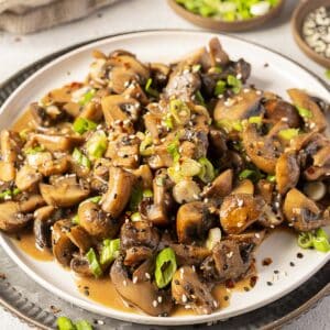 A plate of sautéed mushrooms garnished with chopped green onions and sesame seeds, served on a white dish with additional garnishes in small bowls nearby.
