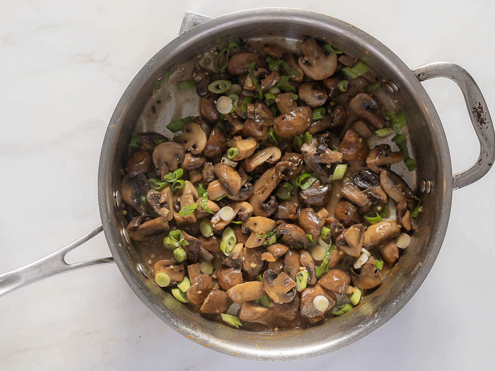 Sliced mushrooms and chopped green onions sautéed in a stainless steel pan on a white surface.