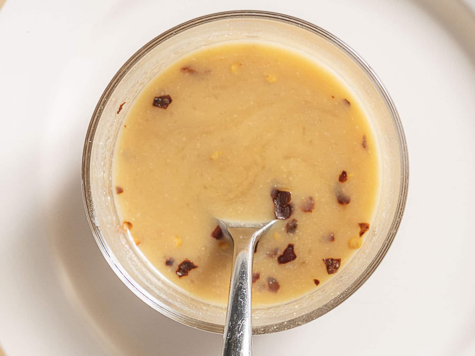 A small glass bowl filled with a light brown creamy sauce, sprinkled with red pepper flakes, with a spoon inside.