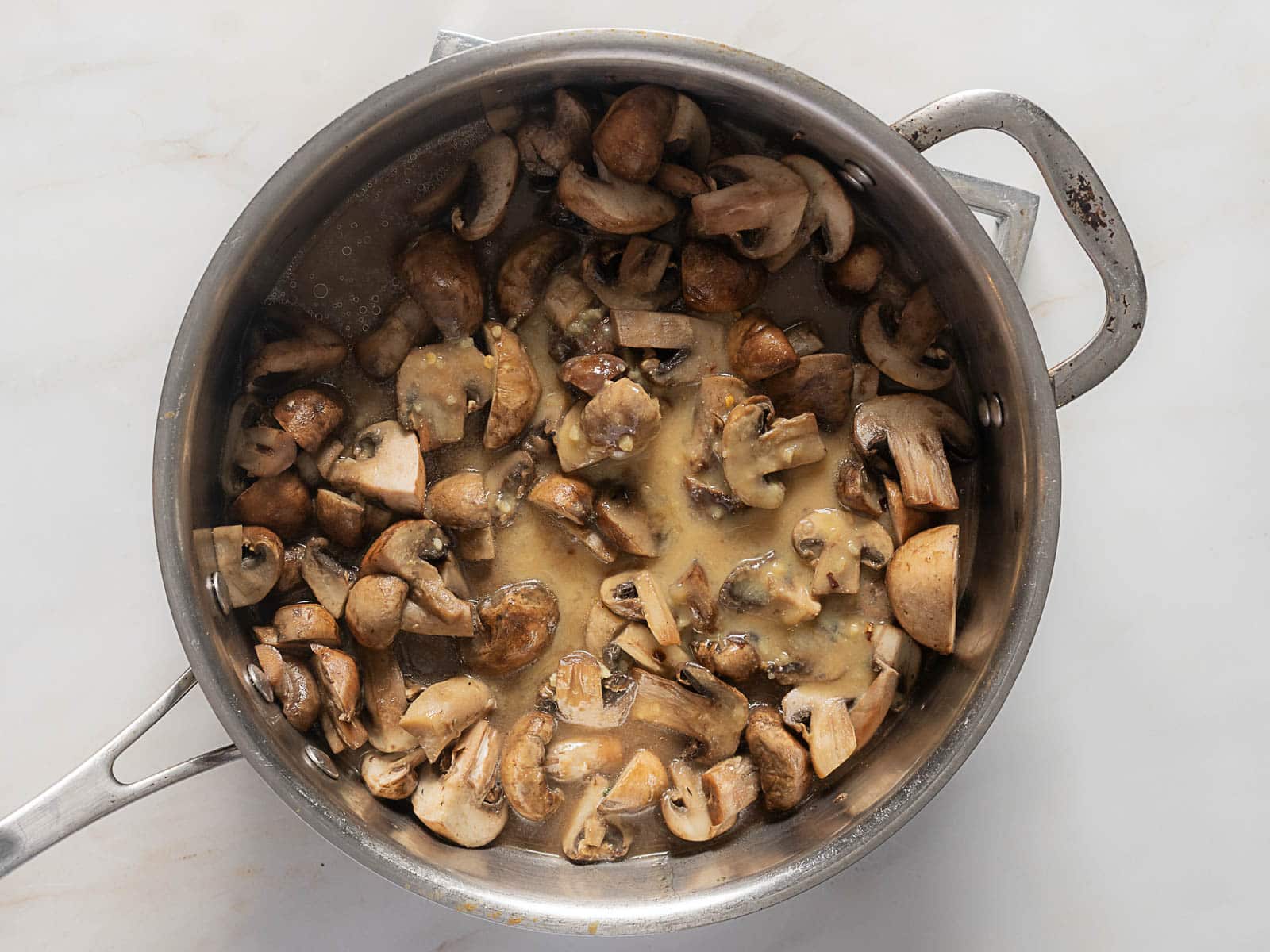 Sliced mushrooms cooking in a stainless steel pan with a light sauce on a white surface.