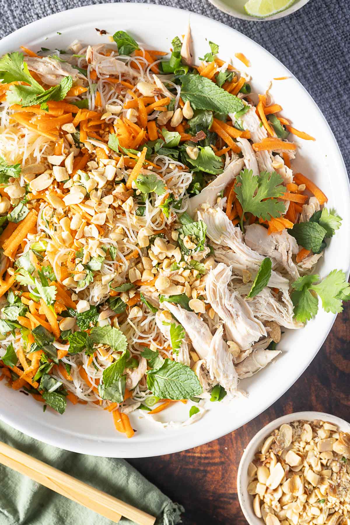A plate of rice noodles with shredded chicken, carrots, peanuts, cilantro, and mint. A small bowl of chopped peanuts and chopsticks are placed beside the plate.