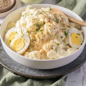 A bowl of potato salad with sliced hard-boiled eggs and a sprinkle of herbs and spices, served with a wooden spoon on a metal tray.
