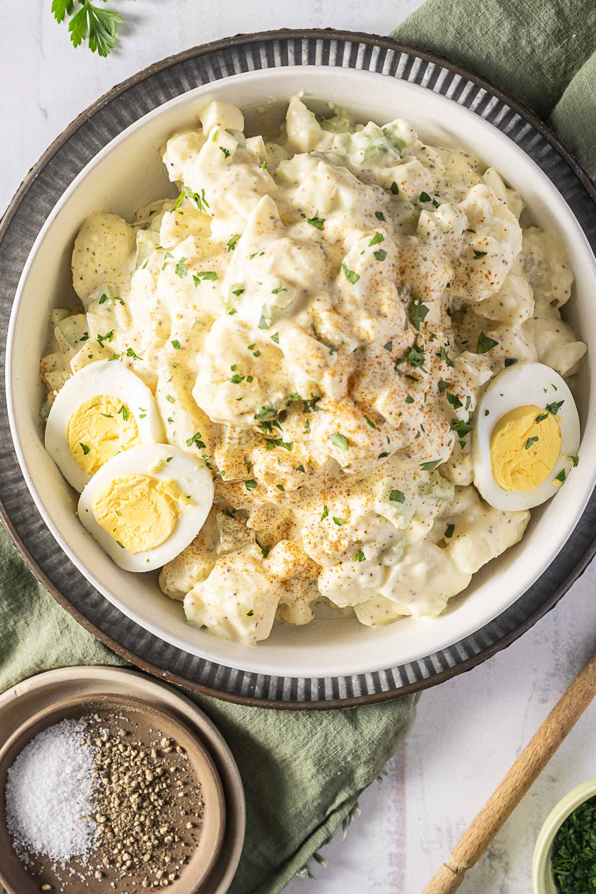 A bowl of creamy potato salad garnished with sliced hard-boiled eggs, sprinkled with parsley and paprika, placed on a green napkin next to small bowls of salt and pepper.