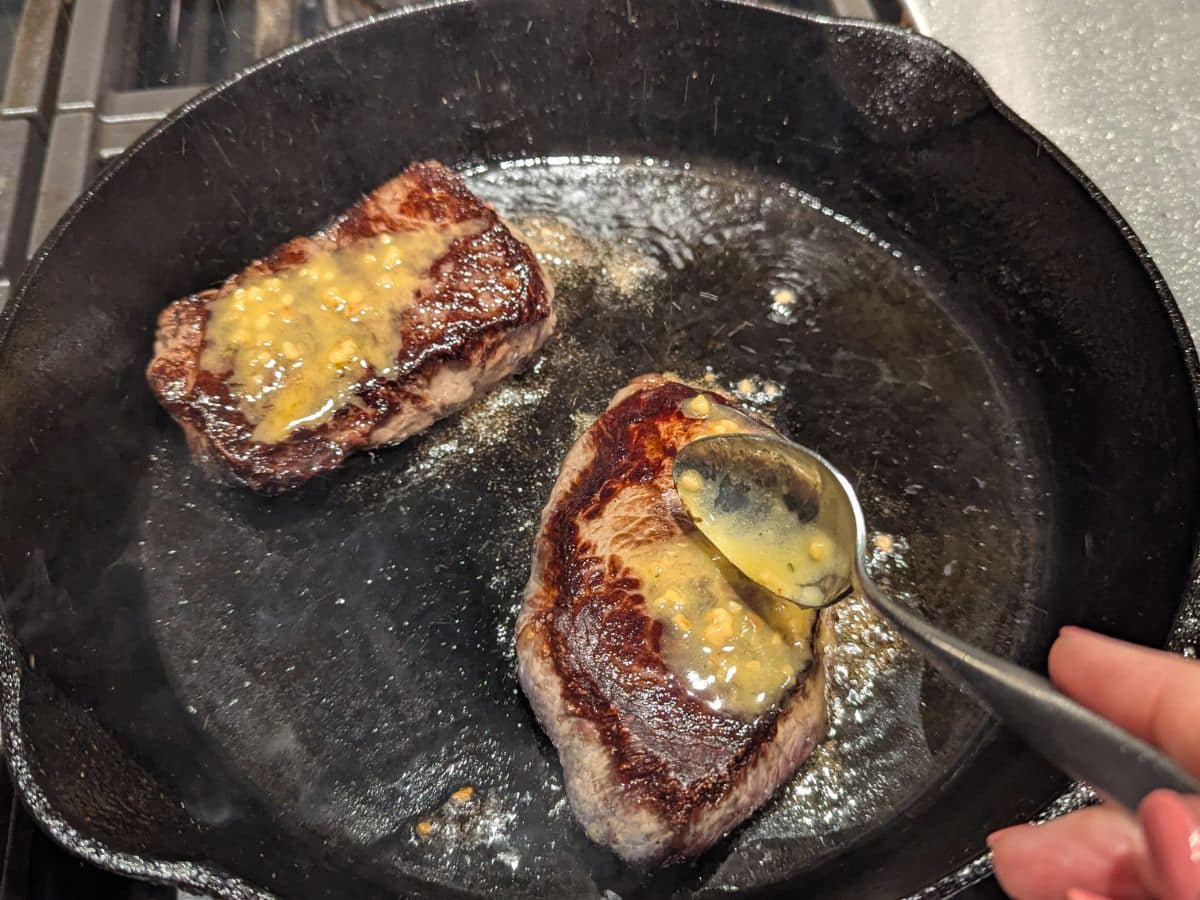 Two steaks cooking in a cast iron skillet, with a person spooning melted garlic butter over them.