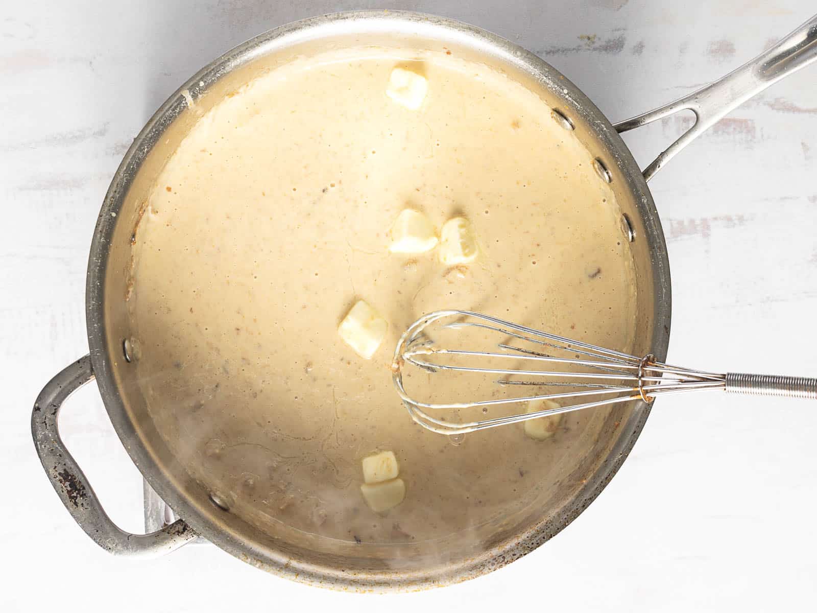 A metal saucepan of creamy sauce with butter cubes on top. A whisk is submerged in the sauce. The pan is on a white surface.