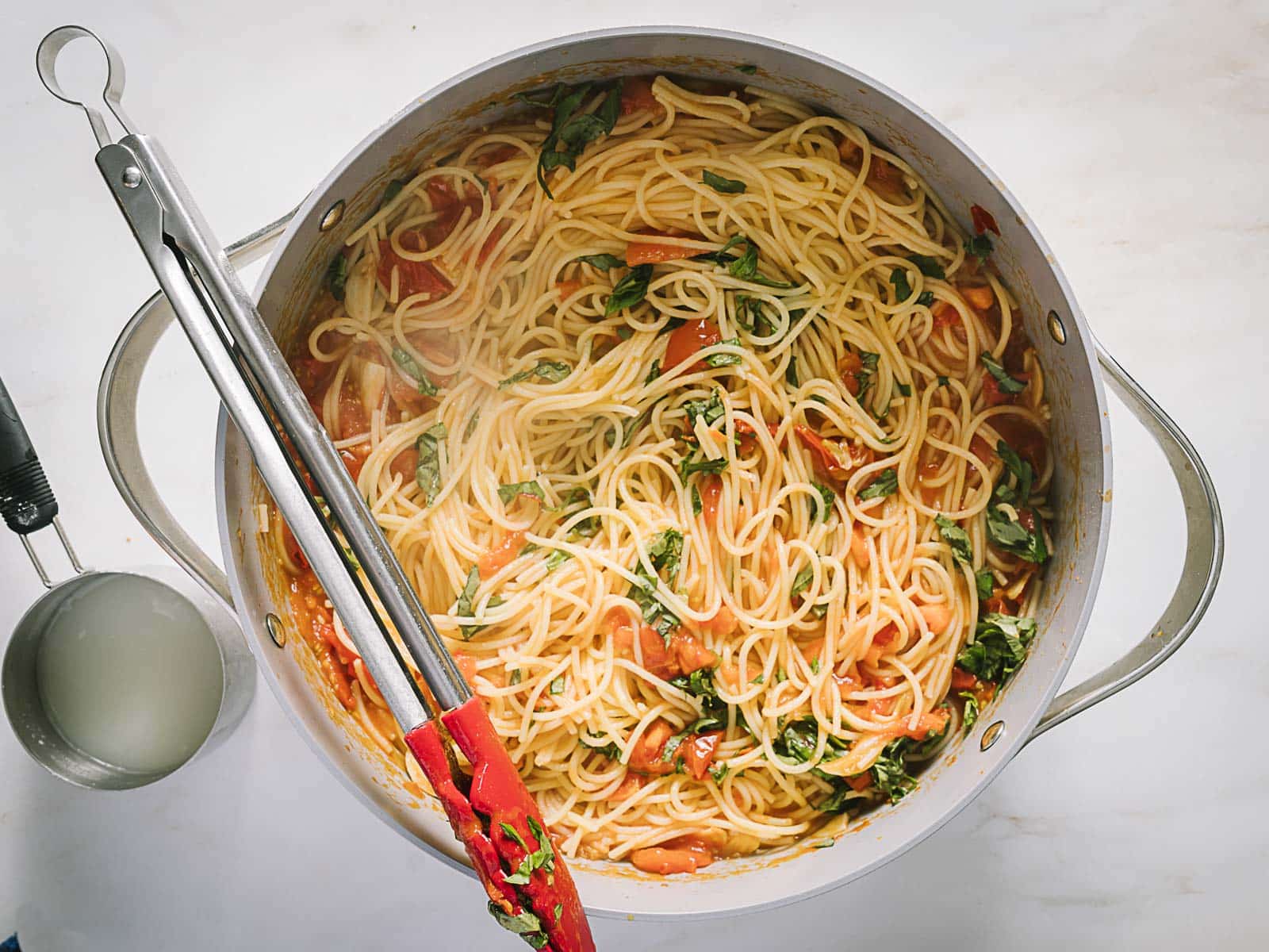A pot of cooked spaghetti mixed with tomato sauce and fresh greens, with metal tongs resting inside and a measuring cup of pasta water nearby.