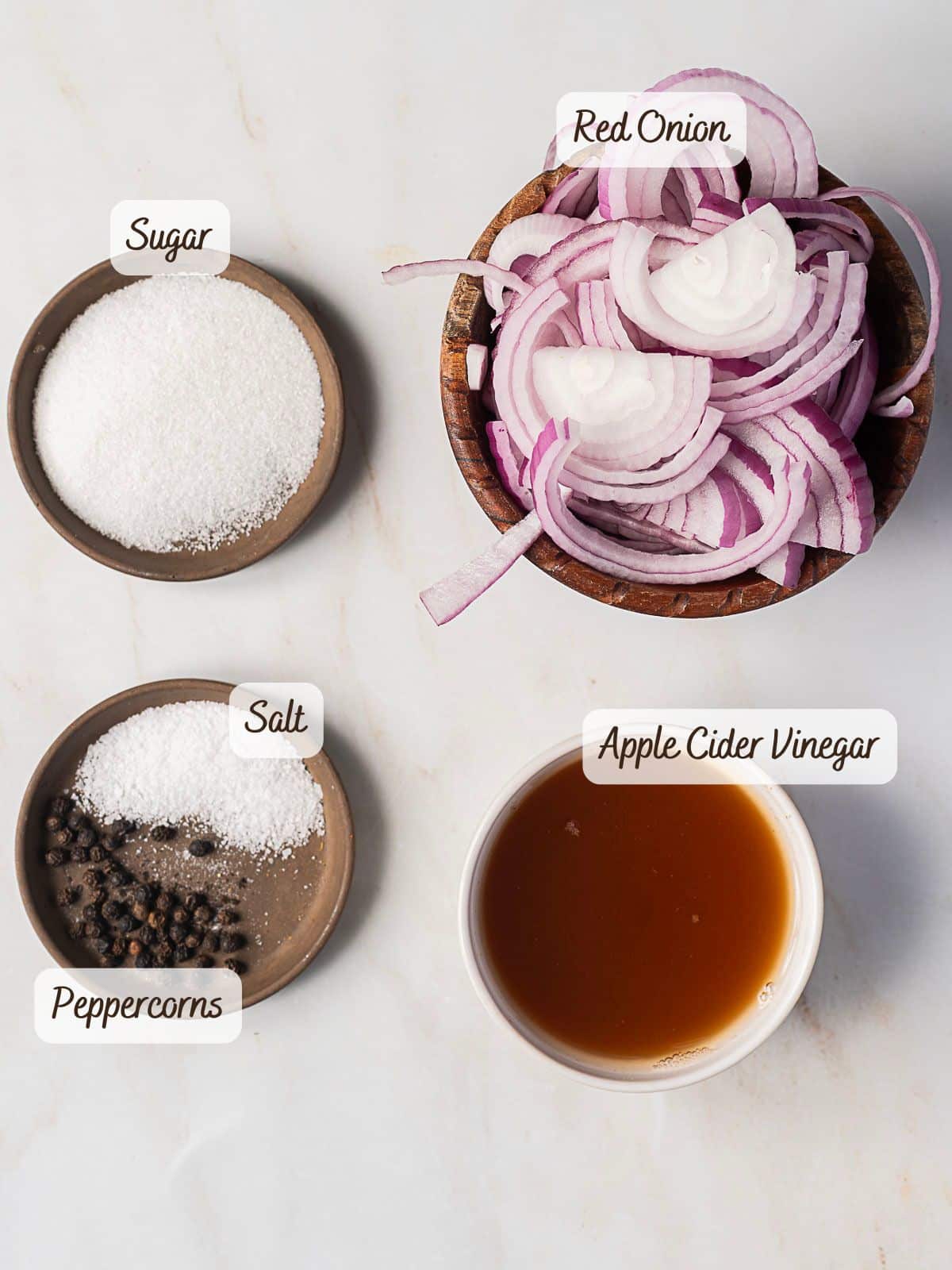 Four labeled ingredients for quick pickled red onions: sliced red onion in a bowl, sugar, salt with peppercorns, and apple cider vinegar in small dishes on a white surface.