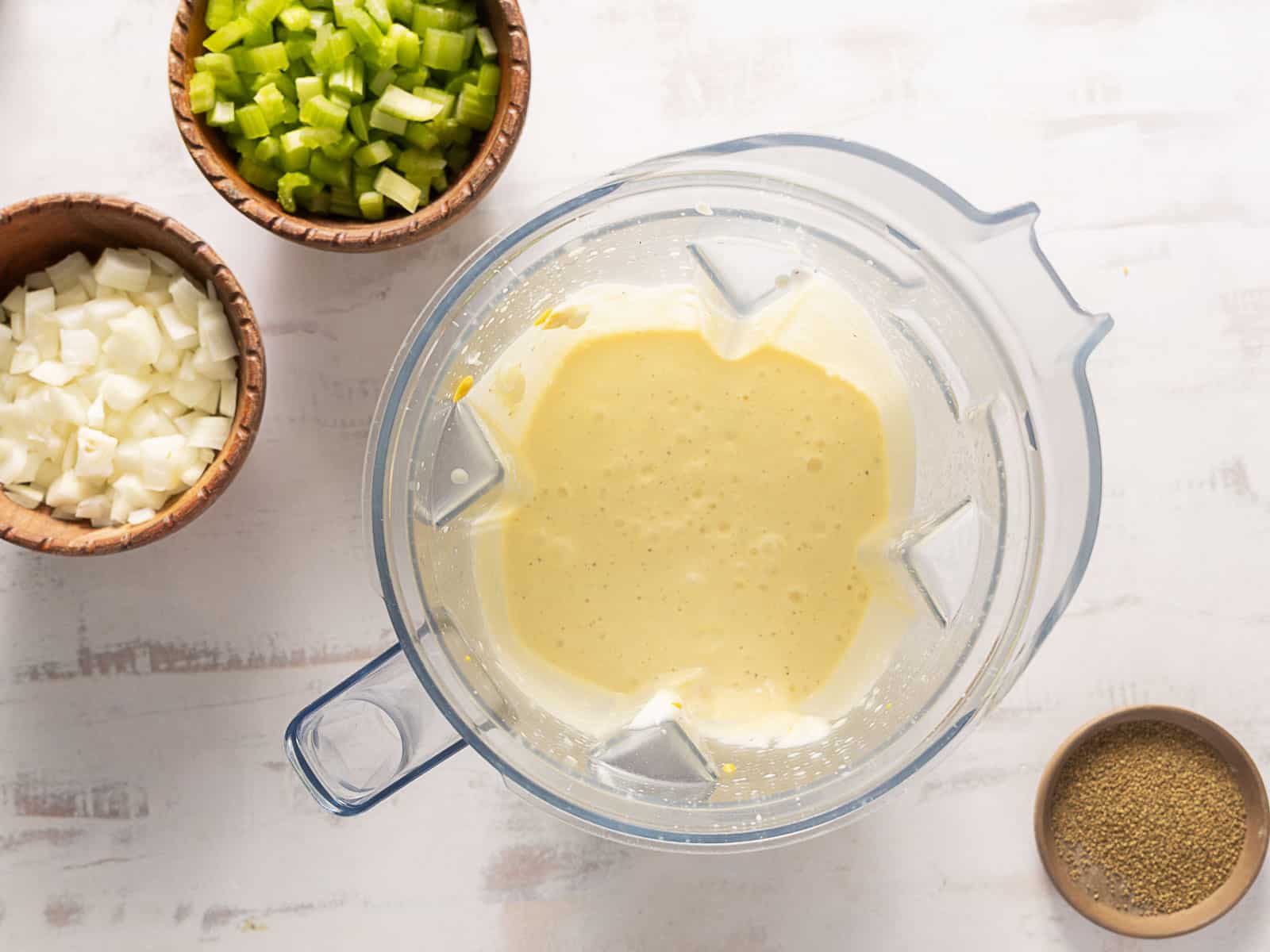 A blender with creamy mixture next to bowls of chopped onions, celery, and a bowl of spices on a white surface.