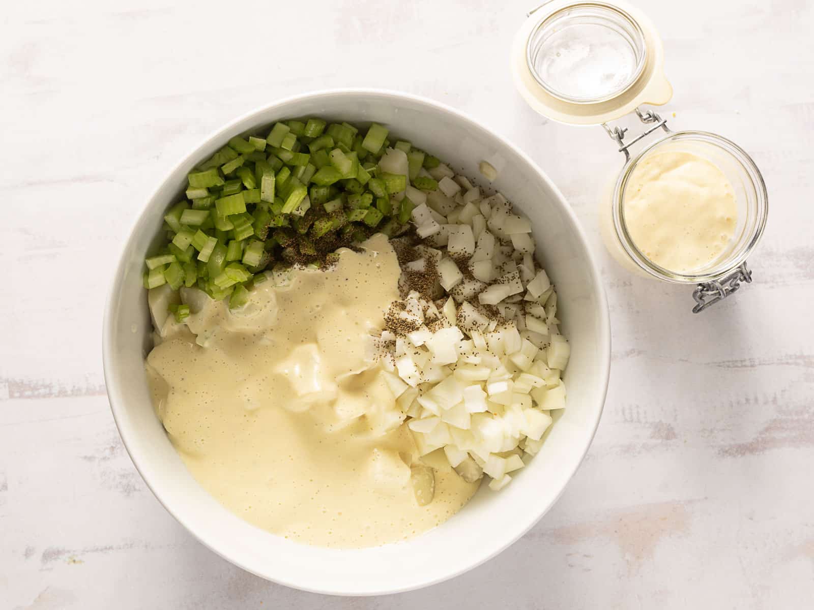 Bowl containing chopped celery, onion, and diced potatoes with a creamy sauce, accompanied by a jar of mayonnaise on the side.
