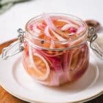 A glass jar filled with sliced red onions soaking in a clear pickling liquid, placed on a white plate.