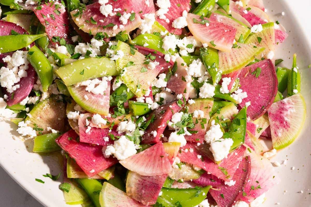 A spotlight-worthy salad featuring sliced watermelon radishes, snow peas, crumbled feta cheese, and parsley on a white plate. Perfect for Easter recipes or any occasion, this vibrant dish is naturally gluten-free and refreshingly delicious.