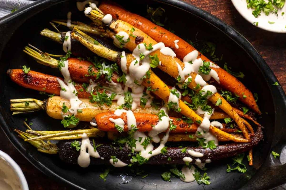 Roasted carrots of various colors take the spotlight on a black platter, drizzled with a creamy sauce and garnished with chopped herbs. Perfect for Easter recipes, this gluten-free dish is both vibrant and delicious.