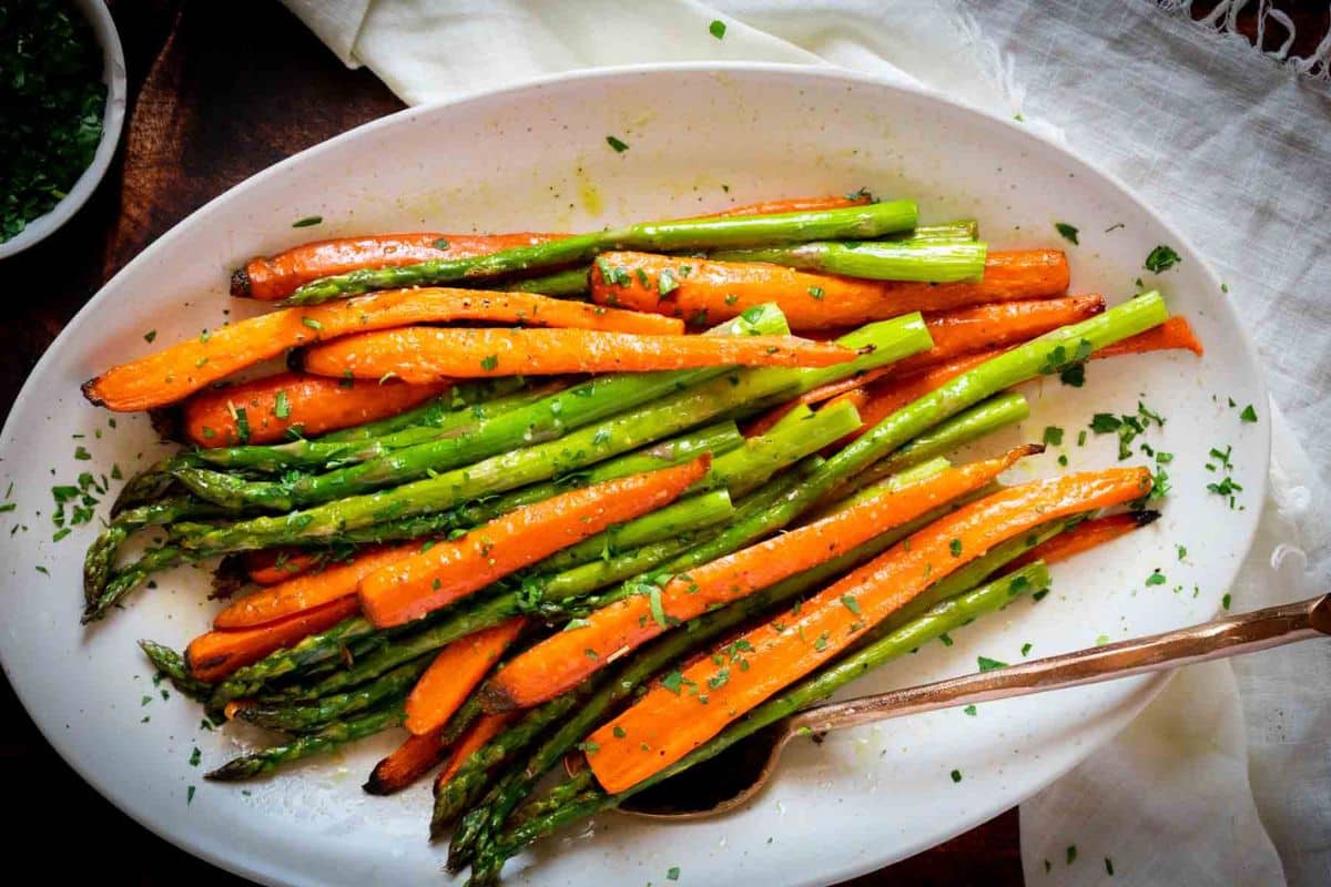 A white plate with cooked carrots and asparagus, garnished with chopped herbs, takes the spotlight in this elegant dish. Perfect for gluten-free Easter recipes, a serving spoon lies invitingly on the side.