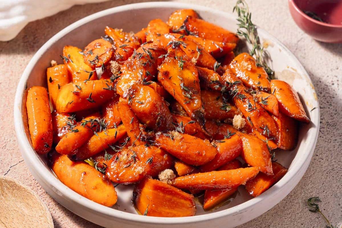 A plate of glazed roasted carrots, perfect for Easter recipes, is garnished with herbs and placed on a textured surface. Gluten-free and delicious!.