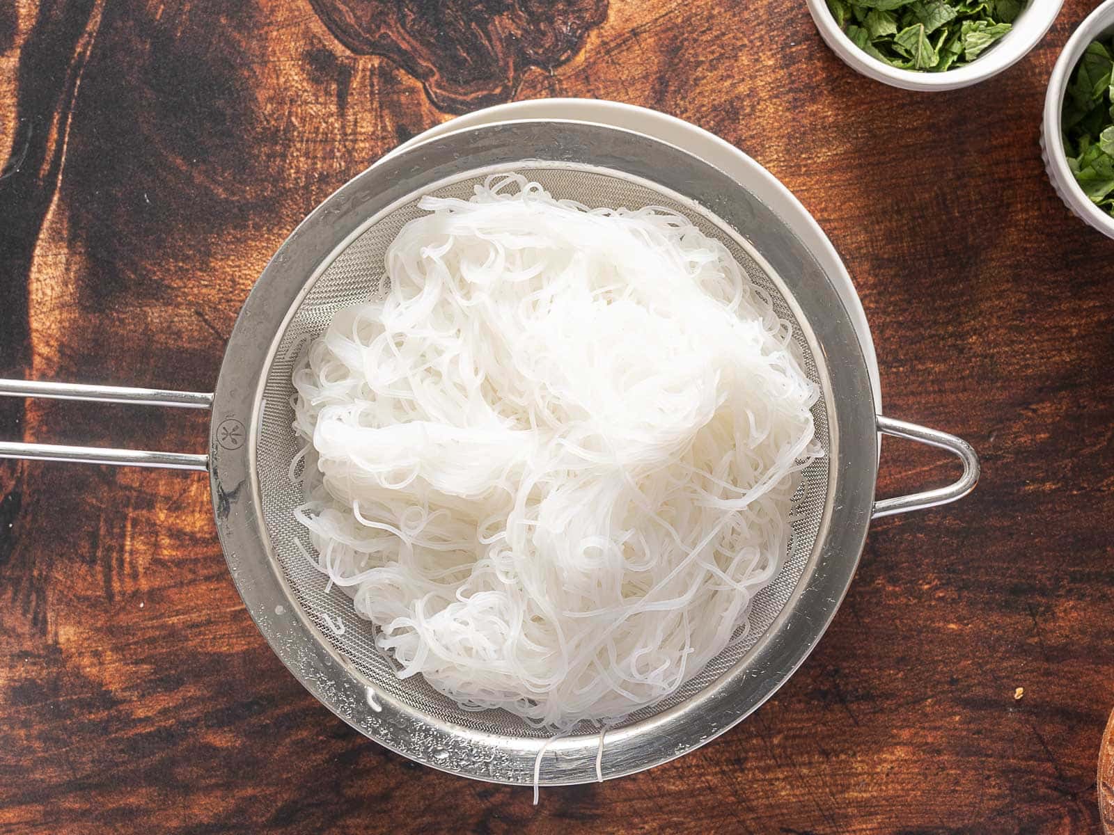 A metal strainer holds cooked rice noodles on a wooden surface, ready to transform into a delightful cold rice noodle salad. Nearby, bowls overflow with fresh herbs and tender chicken, promising a harmonious blend of flavors.