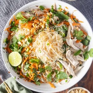 A large white bowl with cold rice noodles, shredded chicken, carrot strips, fresh herbs, crushed peanuts, and a lime wedge rests on a textured gray cloth.