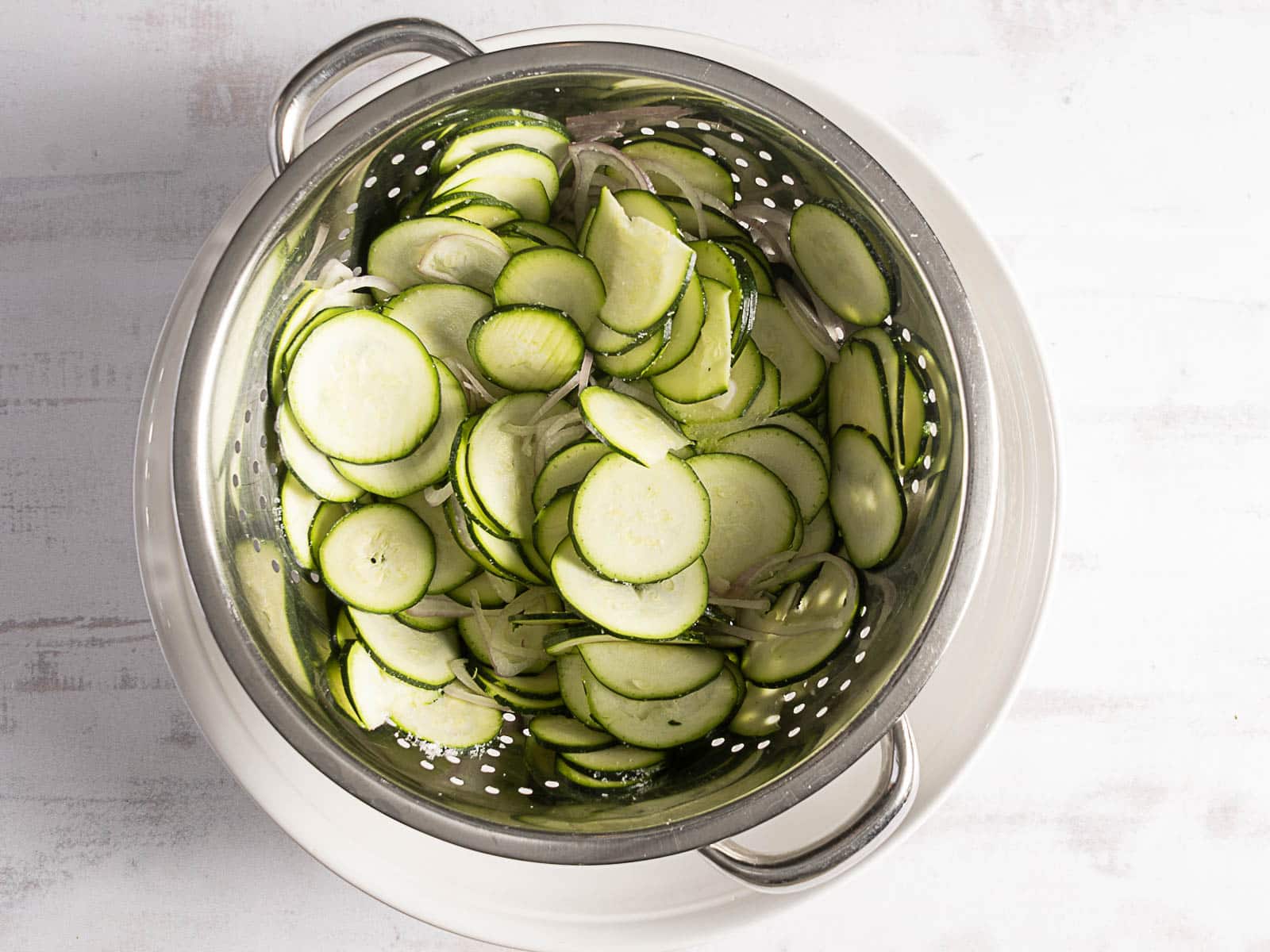 Sliced cucumbers and onions in a metal colander on a white plate.